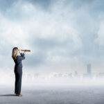 A businesswoman uses a spyglass to look at a city skyline that sits on the distant horizon.