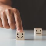3 wooden blocks on a table with smile, straight-line mouth and frown face, with the forefinger of a hand pushing the smiley face forwards