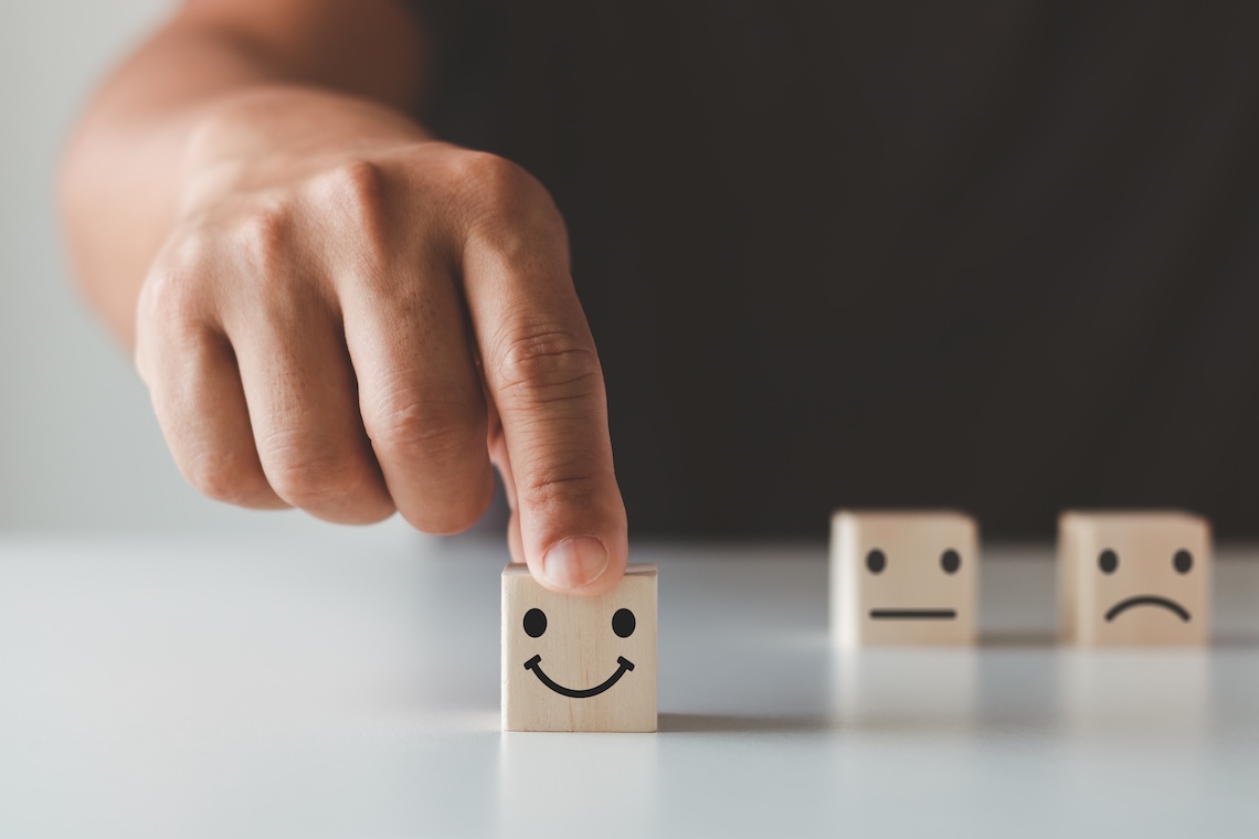 3 wooden blocks on a table with smile, straight-line mouth and frown face, with the forefinger of a hand pushing the smiley face forwards